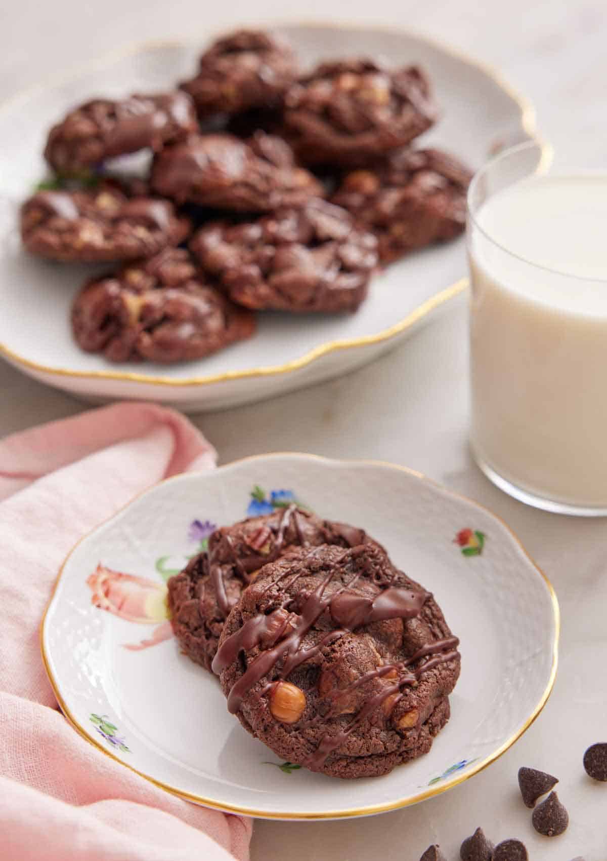 A plate with two turtle cookies with a glass of milk and platter of cookies in the background.