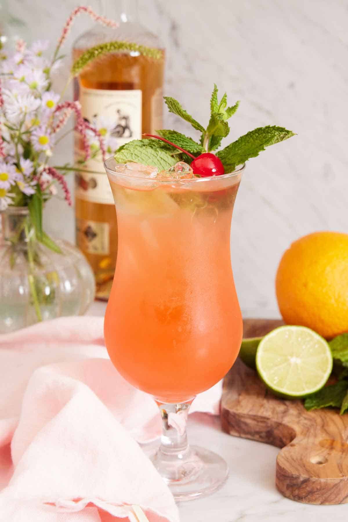 A glass of zombie cocktail garnished with mint leaves and a maraschino cherry. Cut lime and an orange on a cutting board beside it and flowers in the background with a bottle of rum.
