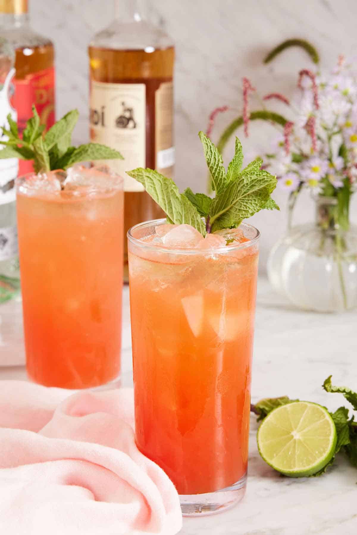 Two glasses of zombie cocktail garnished with mint and a cut lime beside the glasses. Flowers in the background with bottles of alochol.