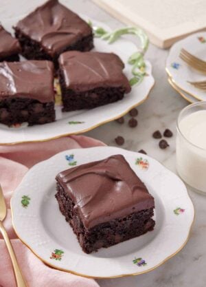A plate with a slice of zucchini brownie with a platter in the background. Glass of milk on the side.
