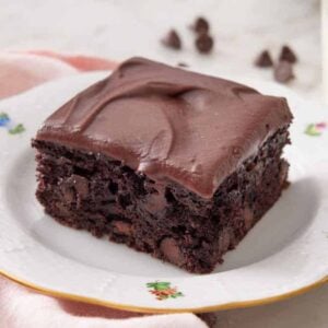 A square piece of zucchini brownie on a plate with a glass of milk and chocolate chips in the background.