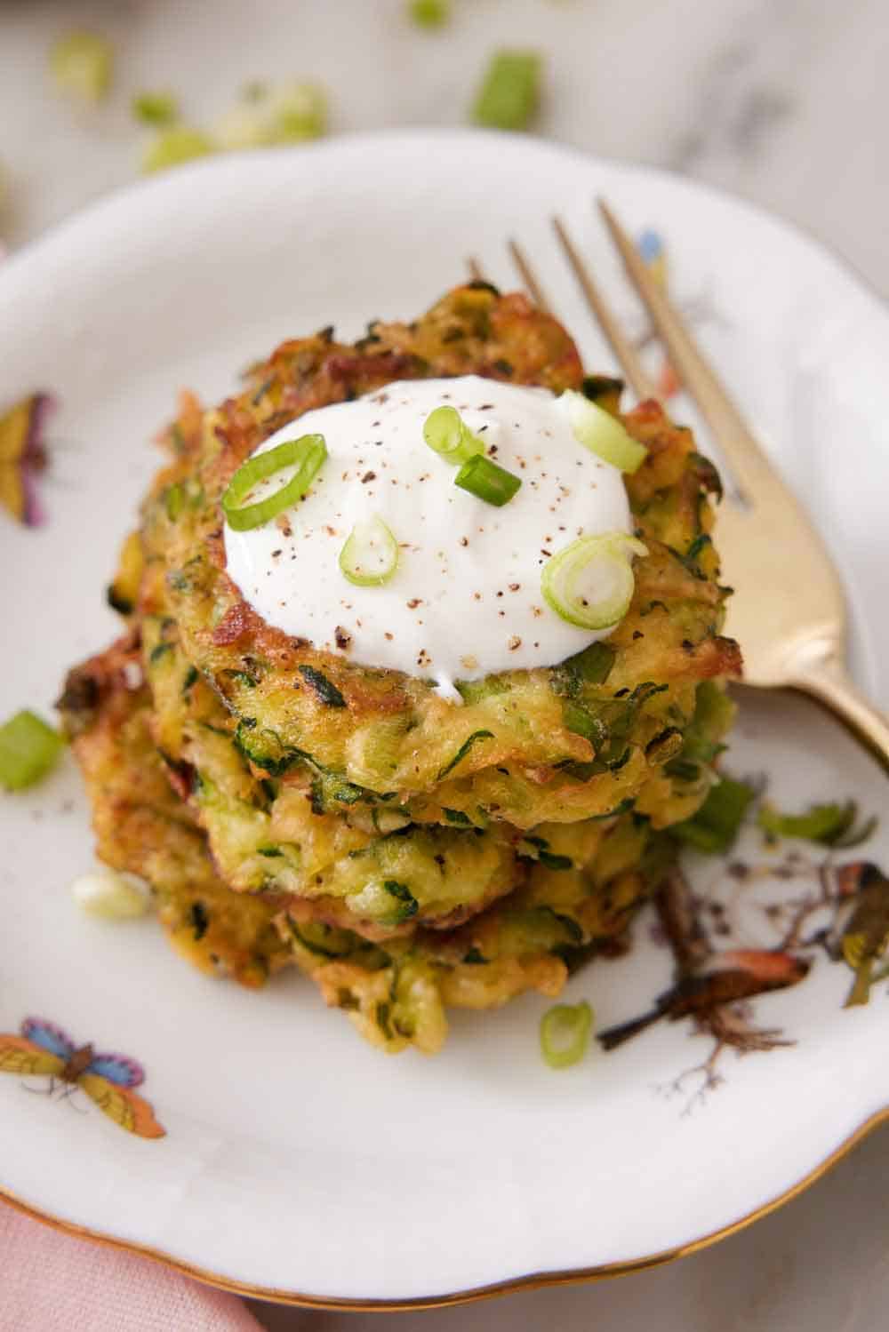 A plate with a stack of zucchini fritters with a dollop of sour cream on top, garnished with green onions.