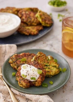 A plate with three zucchini fritters with one with a dollop of sour cream. More zucchini fritters in the background with a glass of iced tea.