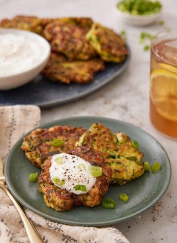 A plate with three zucchini fritters with one with a dollop of sour cream. More zucchini fritters in the background with a glass of iced tea.