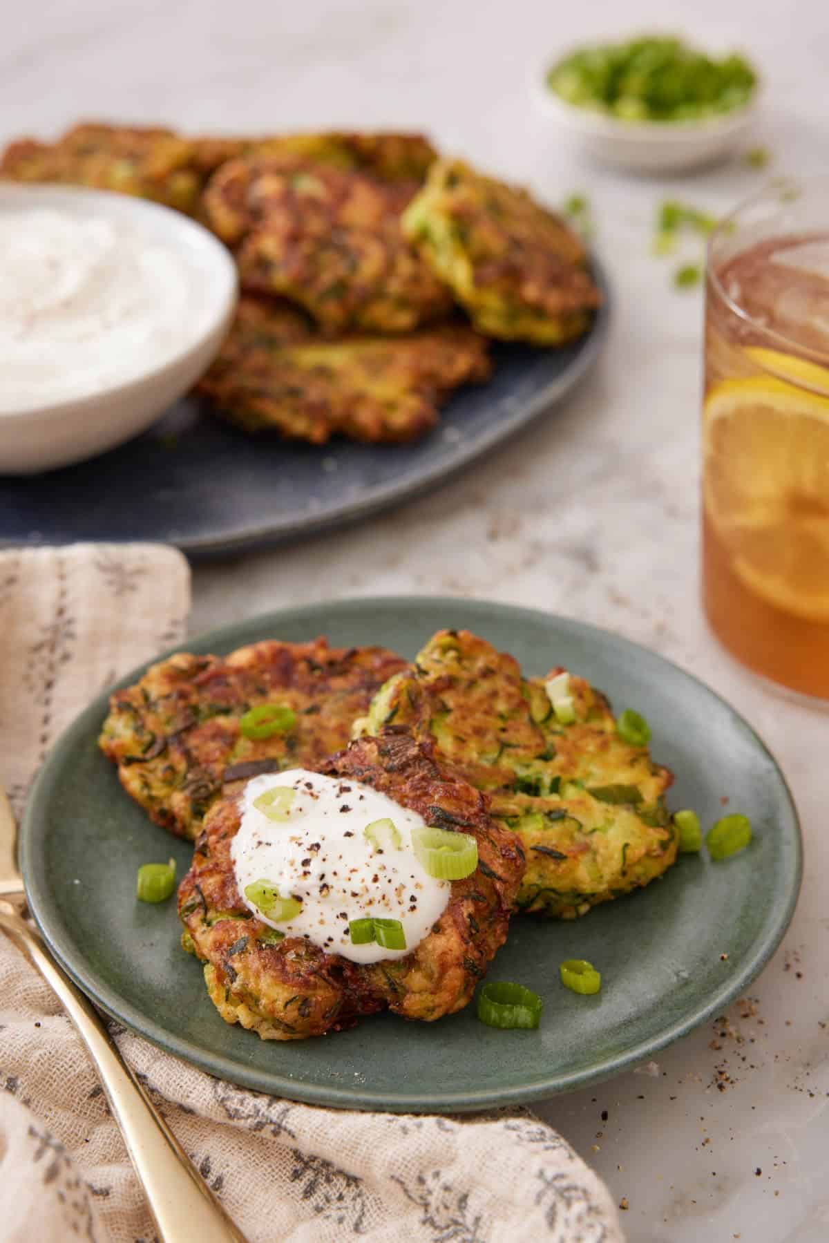 A plate with three zucchini fritters with one with a dollop of sour cream. More zucchini fritters in the background with a glass of iced tea.
