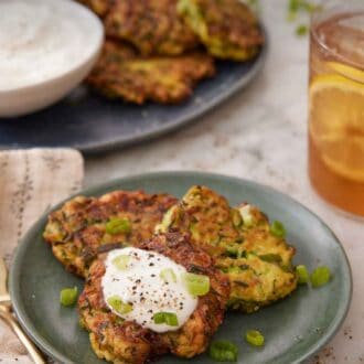 Pinterest graphic of a plate with three zucchini fritters with one with a dollop of sour cream. More zucchini fritters in the background with a glass of iced tea.