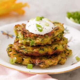 Profile view of a plate of three zucchini fritters with a dollop of sour cream and green onions on top.