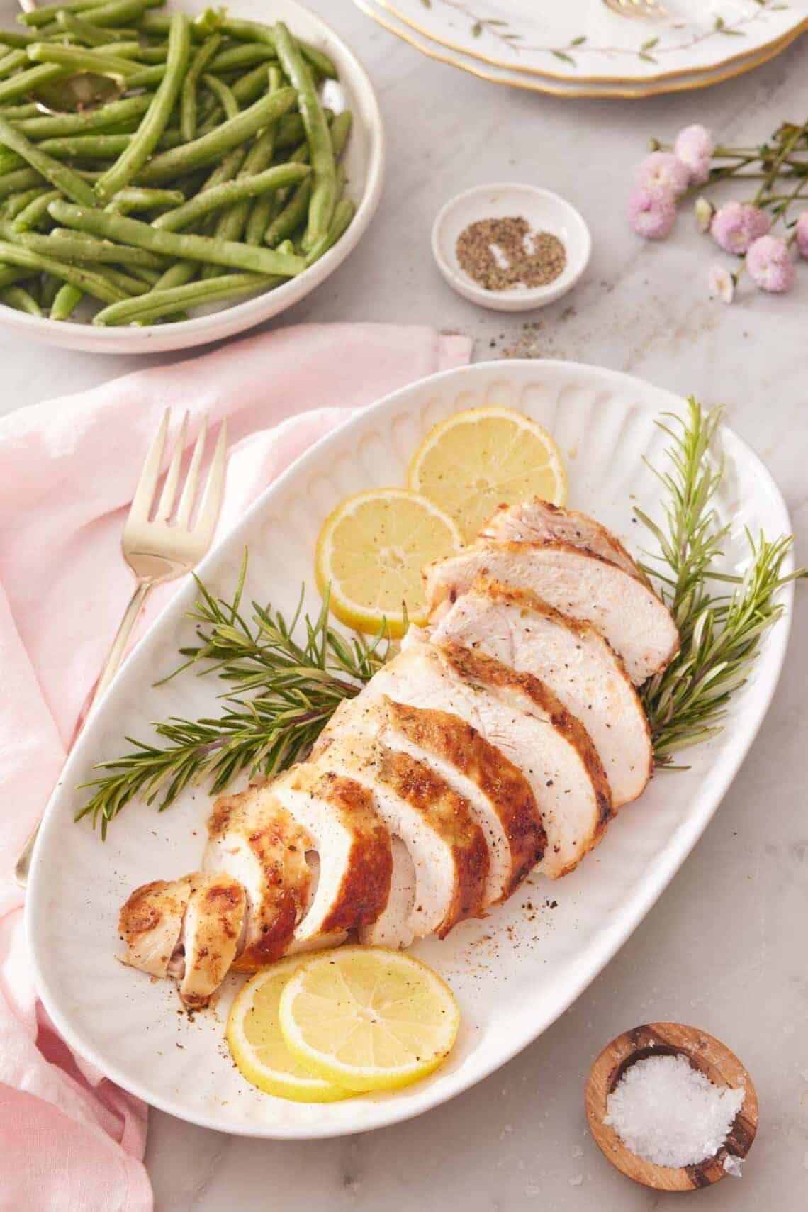 A platter with a sliced air fryer turkey breast on top of rosemary and lemon slices. Green beans in the background.