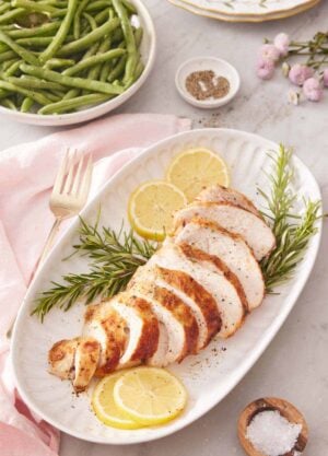 A platter with a sliced air fryer turkey breast on top of rosemary and lemon slices. Green beans in the background.