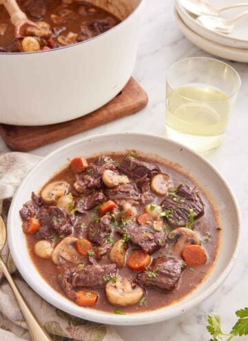 A bowl of beef bourguignon with a glass of wine and pot of more beef bourguignon in the background.