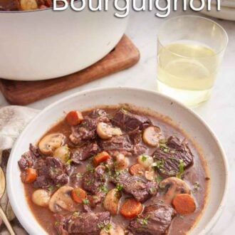 Pinterest graphic of a bowl of beef bourguignon with a glass of wine and pot of more beef bourguignon in the background.