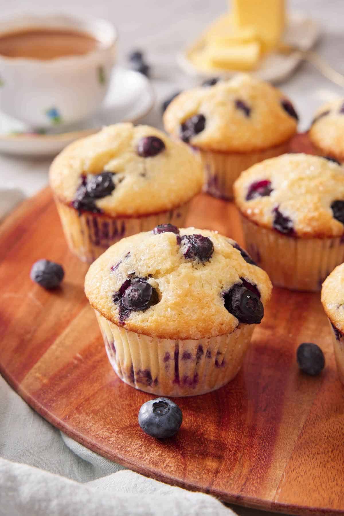 A round wooden serving board with multiple blueberry muffins with some blueberries scattered around.
