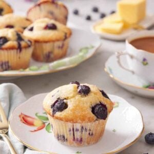 A plate with a blueberry muffin with additional muffins in the background along with a cup of coffee and butter.