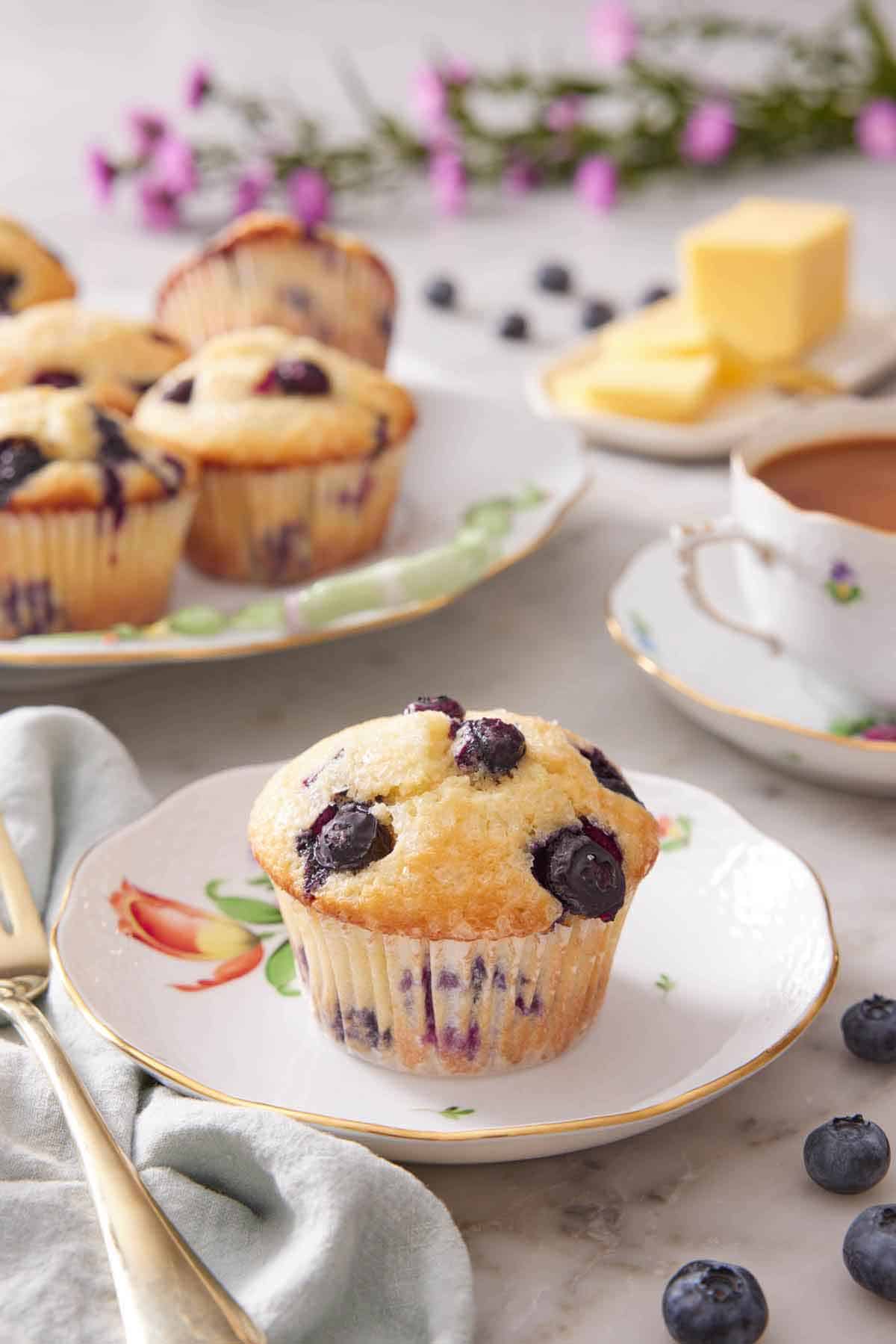 A plate with a blueberry muffin with additional muffins in the background along with a cup of coffee and butter.