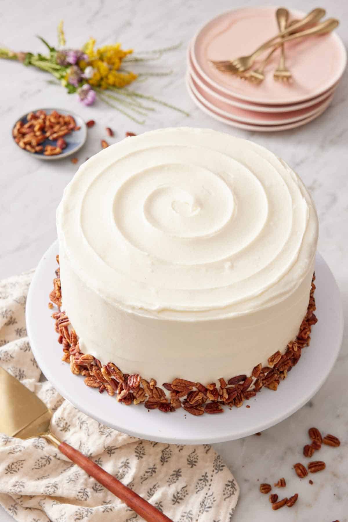 A butter pecan cake on a cake stand. A stack of plates and forks in the background with a bowl of pecans and some flowers.