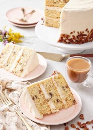 Two plated slices of butter pecan cake with the rest of the cake on a cake stand in the background with a glass of coffee.