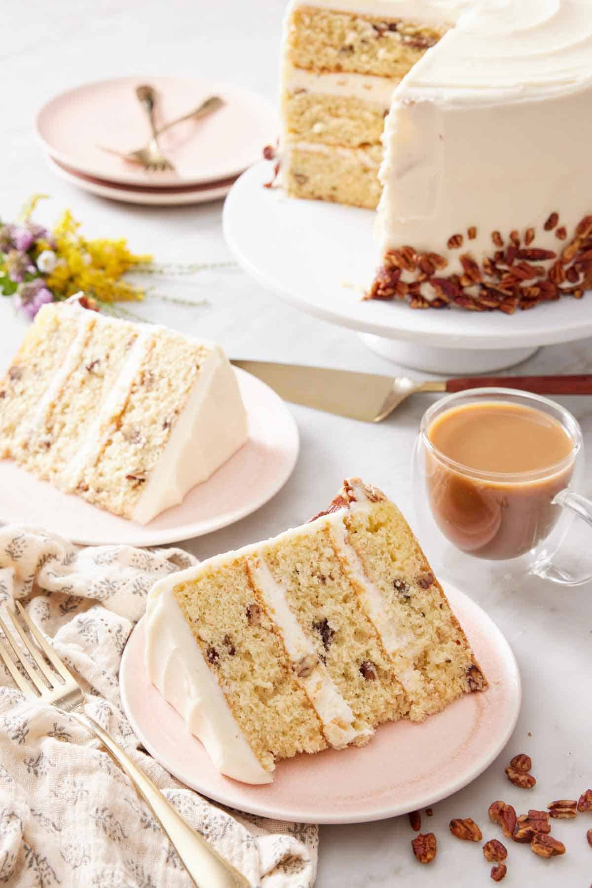 Two plated slices of butter pecan cake with the rest of the cake on a cake stand in the background with a glass of coffee.