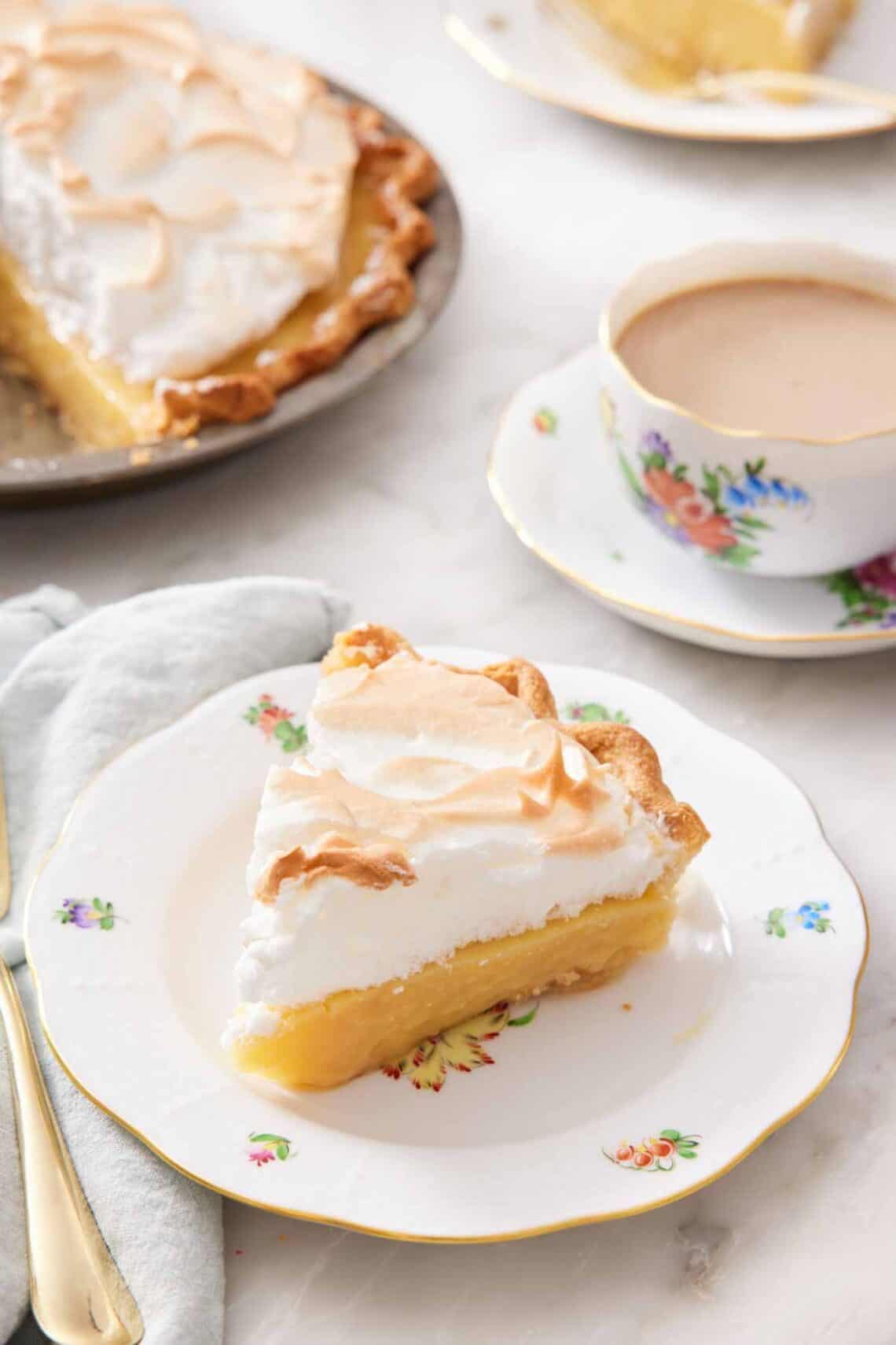 A plate with a slice of butterscotch pie with a cup of coffee in the background and the rest of the pie.