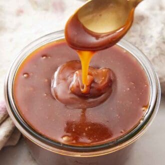 A jar of caramel sauce with a spoon lifted up from it, drizzling the sauce down back into the jar.
