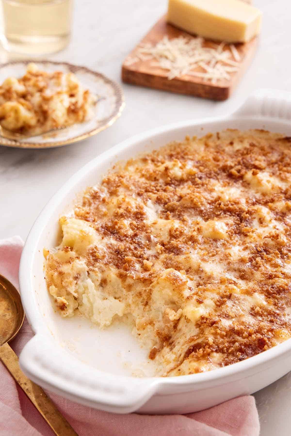 A white baking dish of cauliflower gratin with a serving scooped out onto the plate in the background. Shredded cheese on a board in the back.