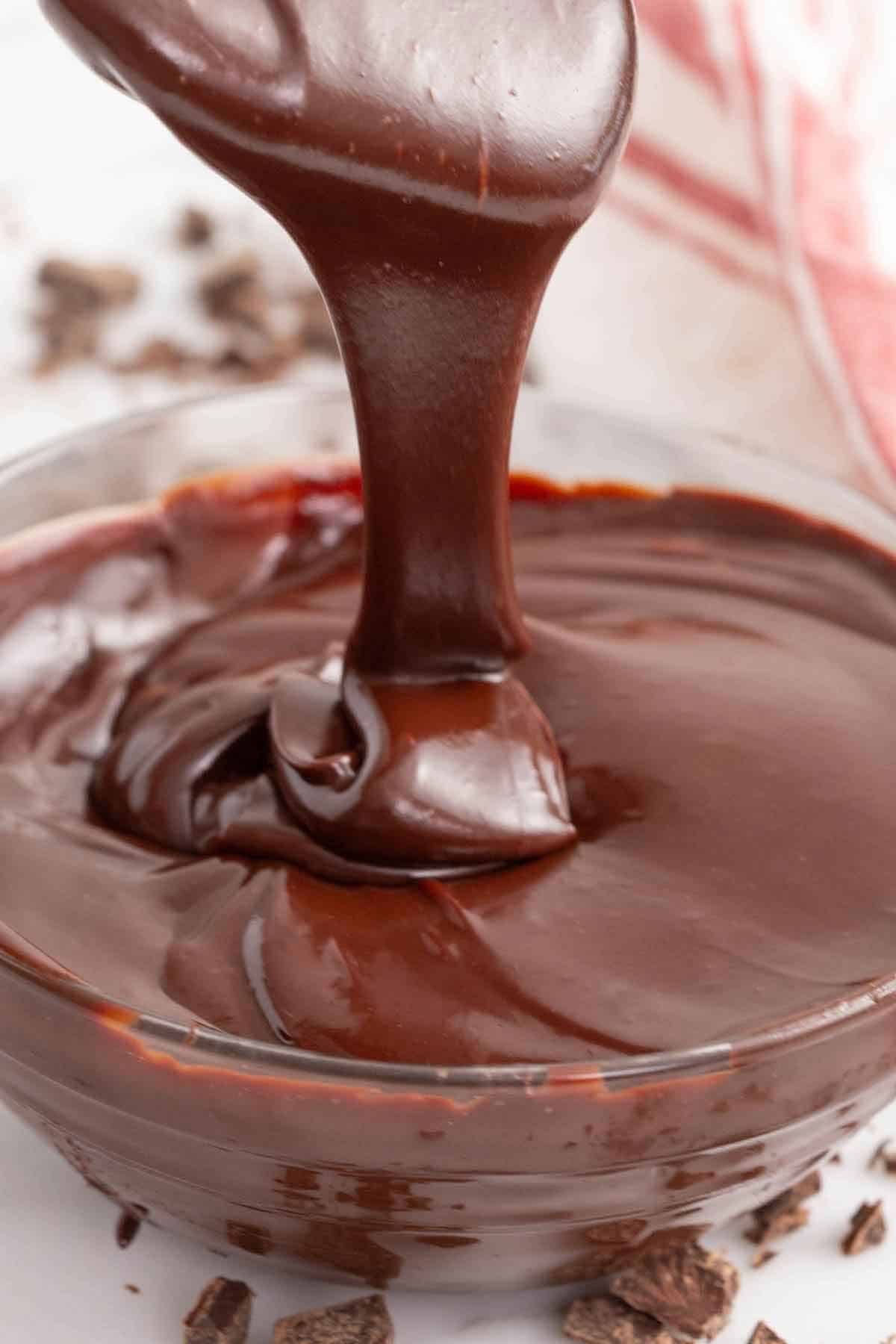 A glass bowl of chocolate ganache with a spatula lifted up with ganache drizzling off.