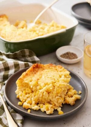 A plate with a serving of corn casserole with a baking dish, pepper, and drink in the background.