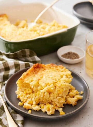 A plate with a serving of corn casserole with a baking dish, pepper, and drink in the background.