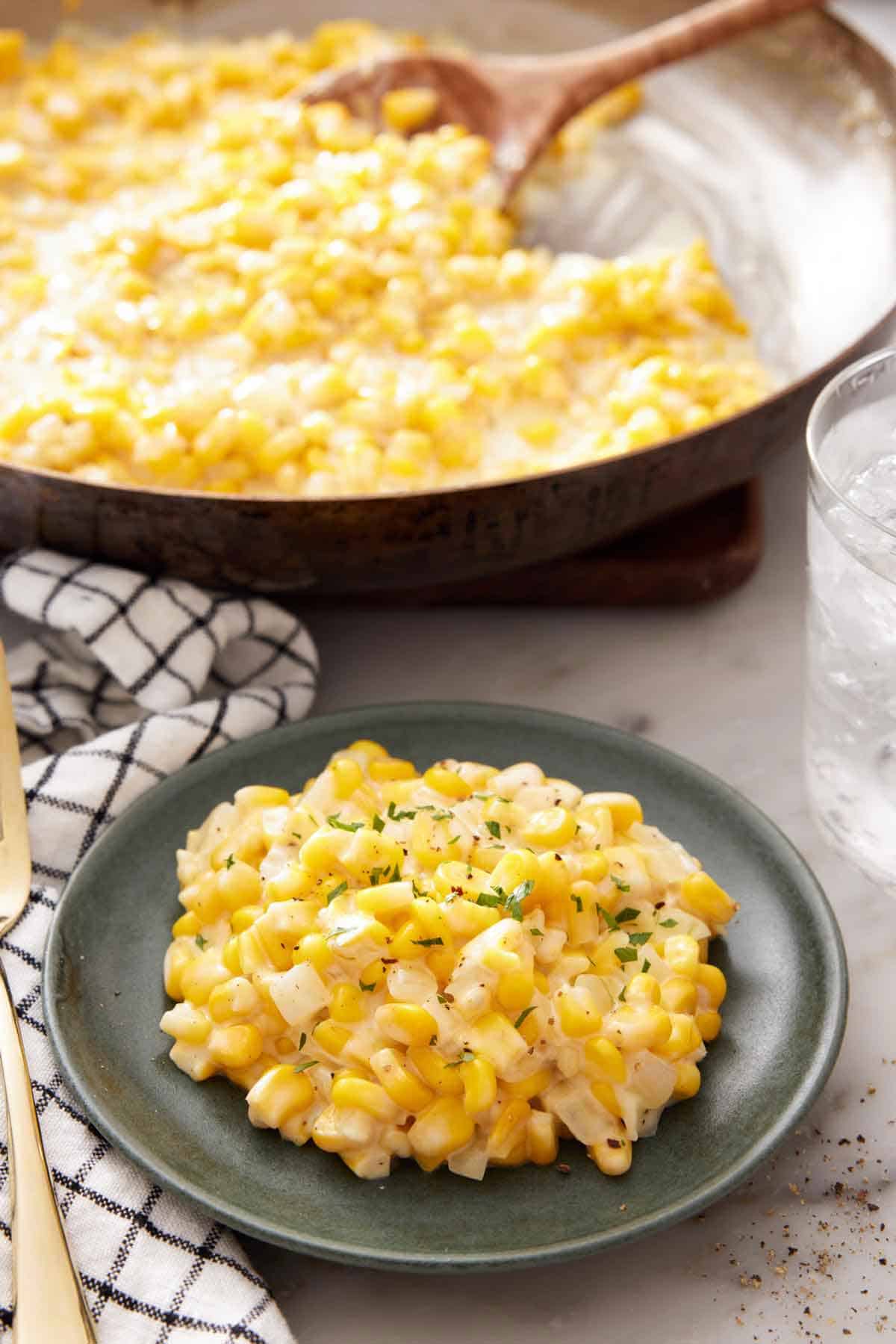 A plate of creamed corn with a skillet with more creamed corn and a wooden spoon in the background.