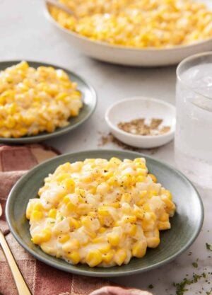 Two plates of creamed corn with a platter with more in the background along with a glass of water and bowl of pepper.