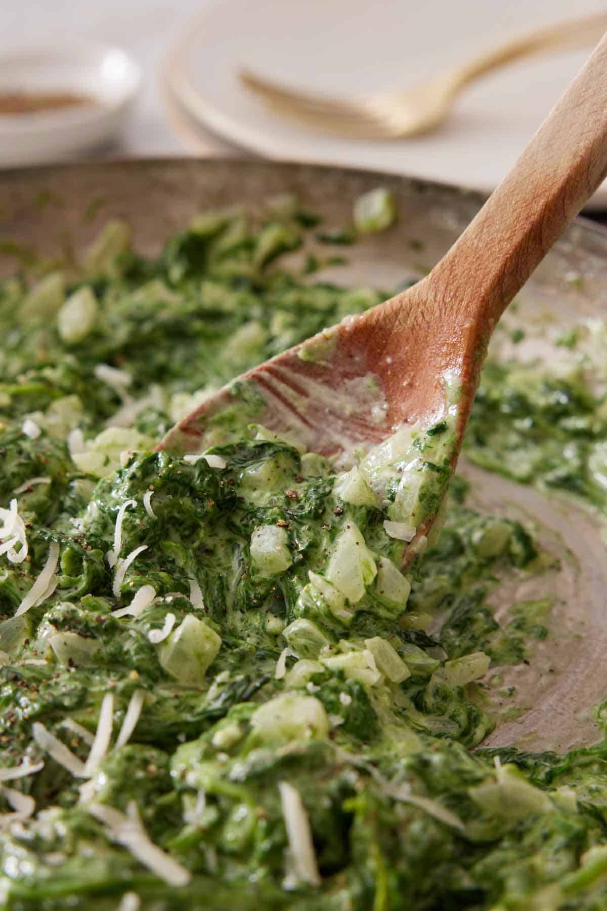 A wooden spoon stirring creamed spinach in a skillet.