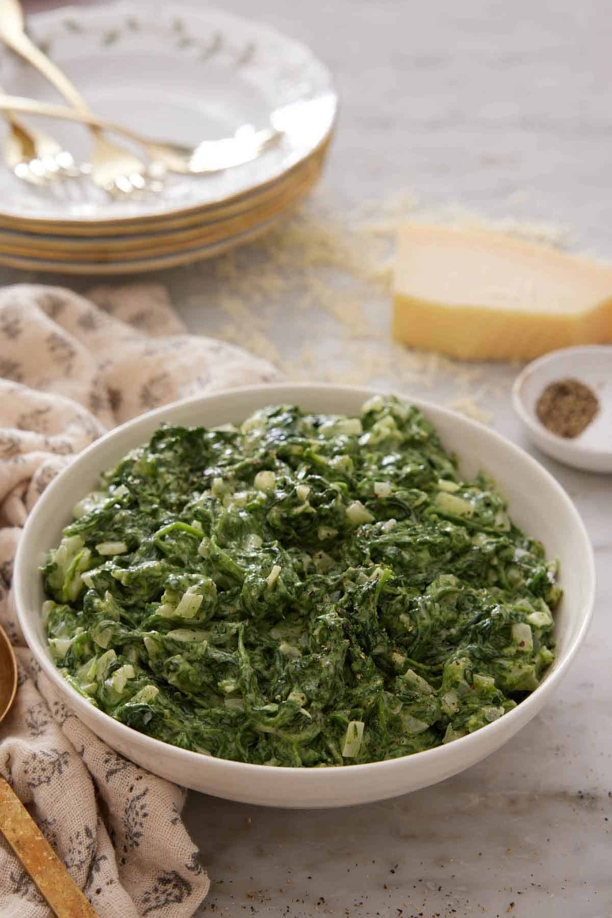 A bowl of creamed spinach with a block of grated parmesan, stack of plates, and forks in the background.