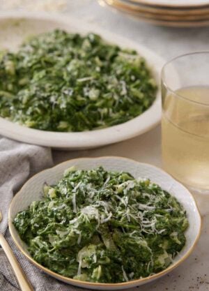 A bowl of creamed spinach with a drink and serving platter of creamed spinach in the background.