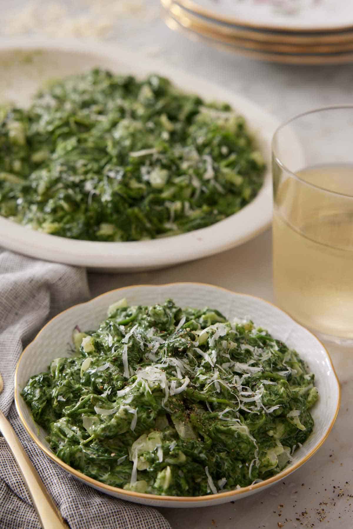 A bowl of creamed spinach with a drink and serving platter of creamed spinach in the background.