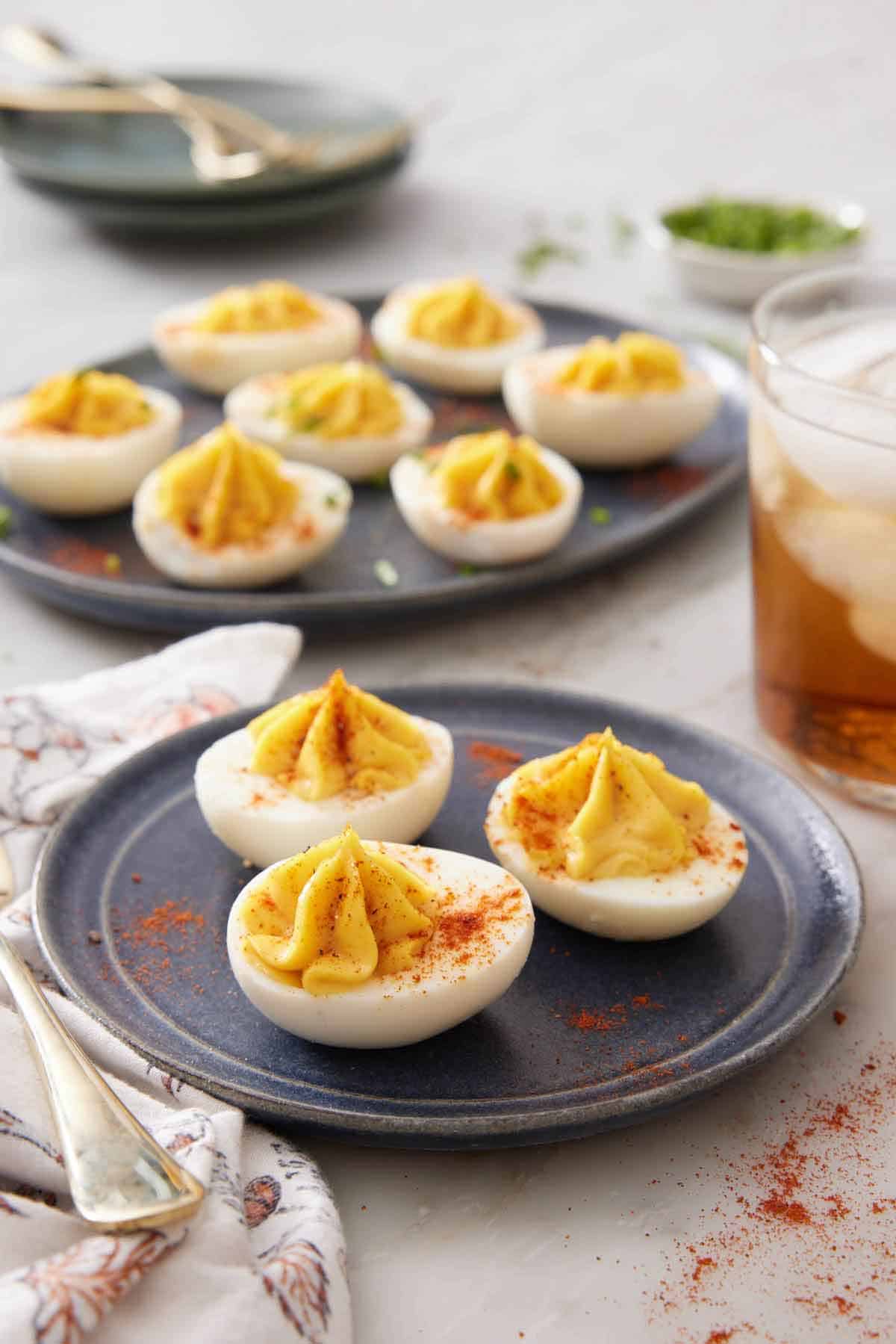 A plate with three deviled eggs with an additional platter in the background with more. A drink off to the side and paprika sprinkled around.