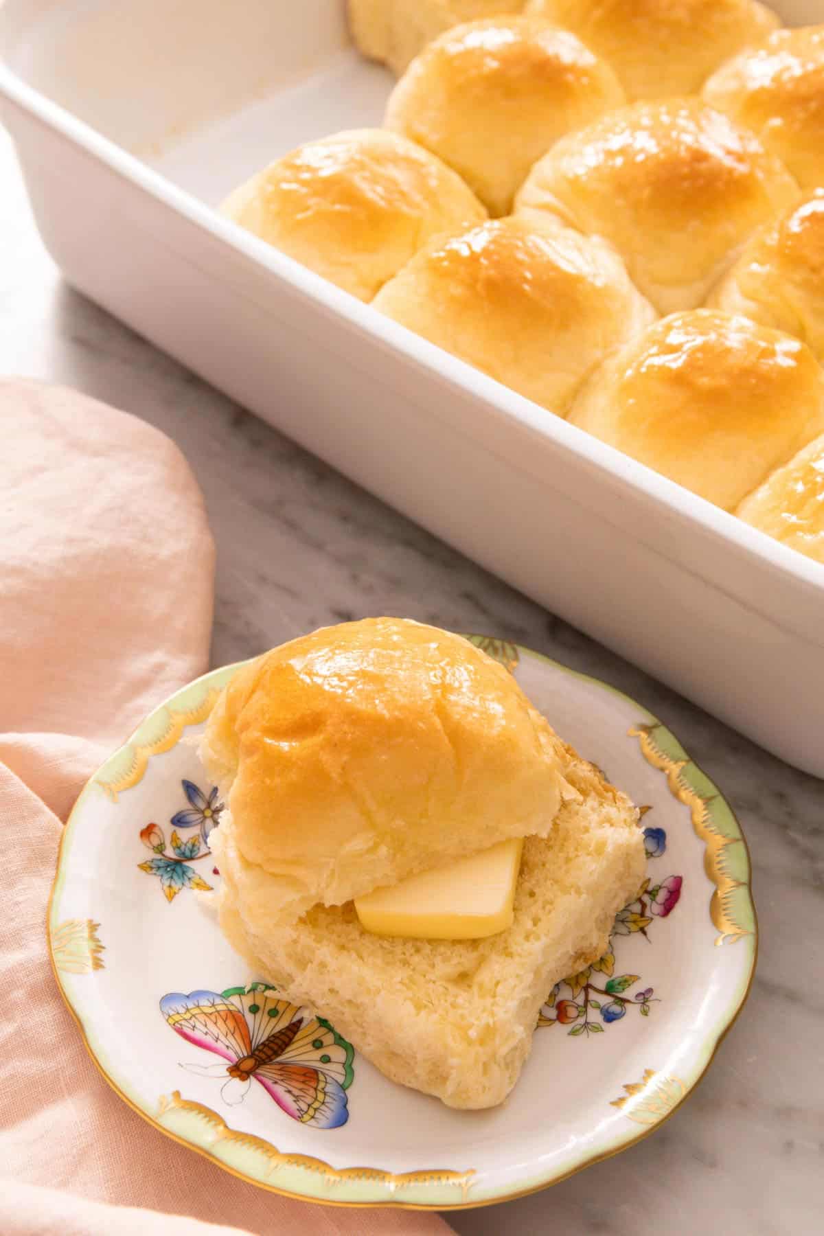 A plate with a dinner roll, sliced, with a piece of butter placed in between. A baking dish with more rolls in the background.