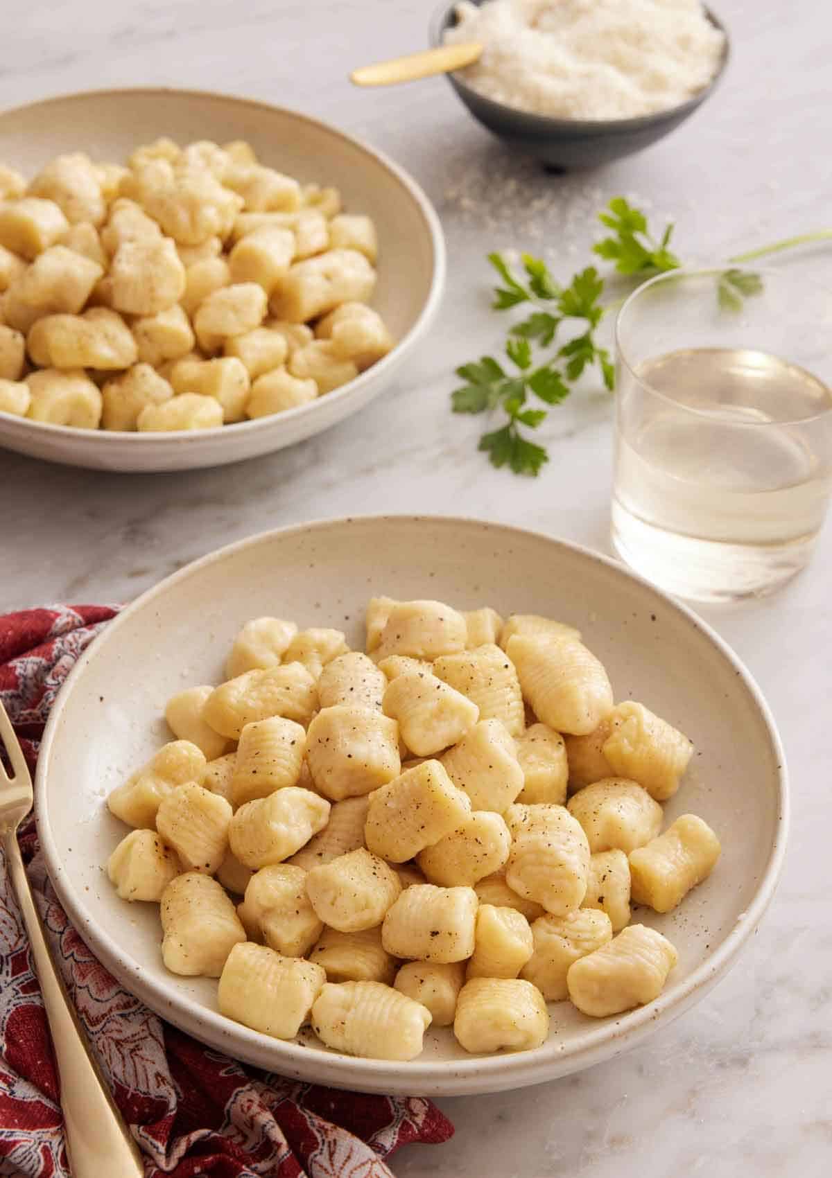 Two plates of gnocchi with a glass of wine and bowl of cheese.