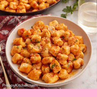 Pinterest graphic of a skillet and plate of gnocchi coated in a red sauce. Glass of wine and bowl of shredded cheese in the background.
