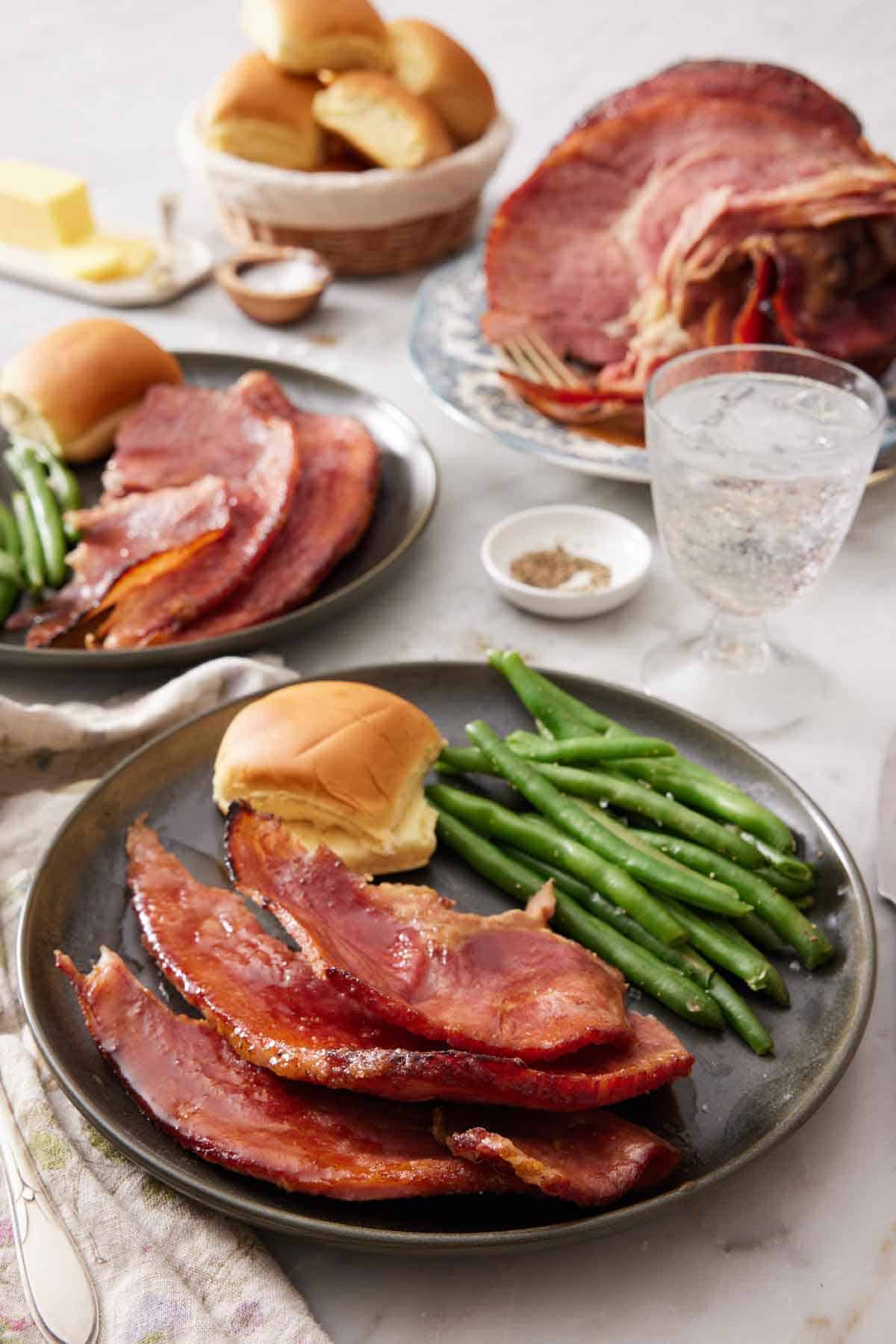 A plate with a serving of honey baked ham with bread and green beans. A second plated serving in the background along with a platter with the ham. A glass of water and bowl of bread in the back.
