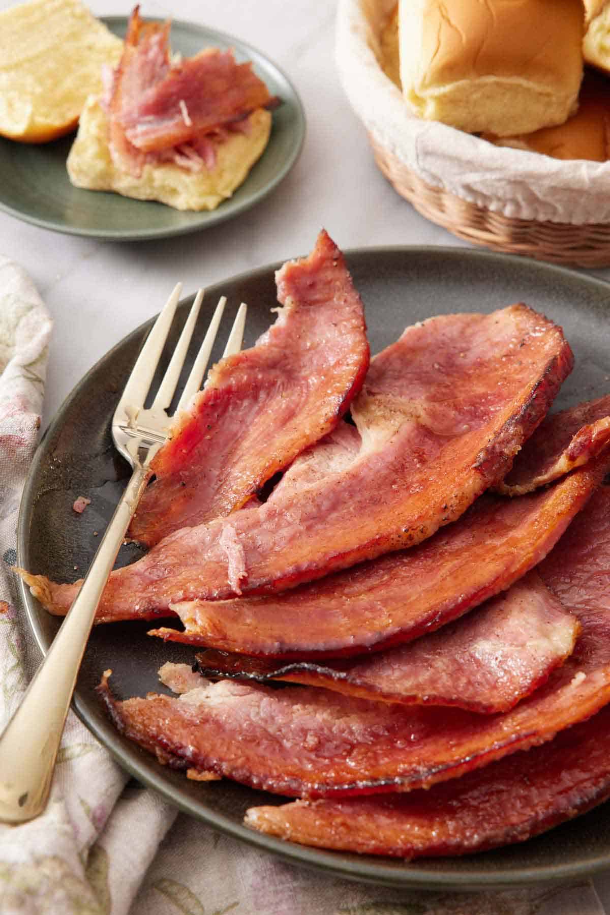 A plate with a pieces of honey baked ham with a fork. A plate with bread and ham on it in the background.