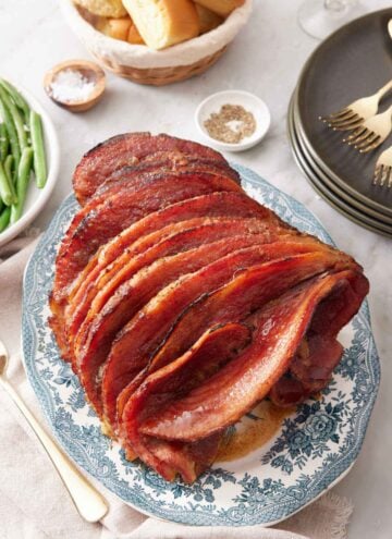 A platter with a honey baked ham with a stack of plates, bowl of bread, and some green beans off to the side.