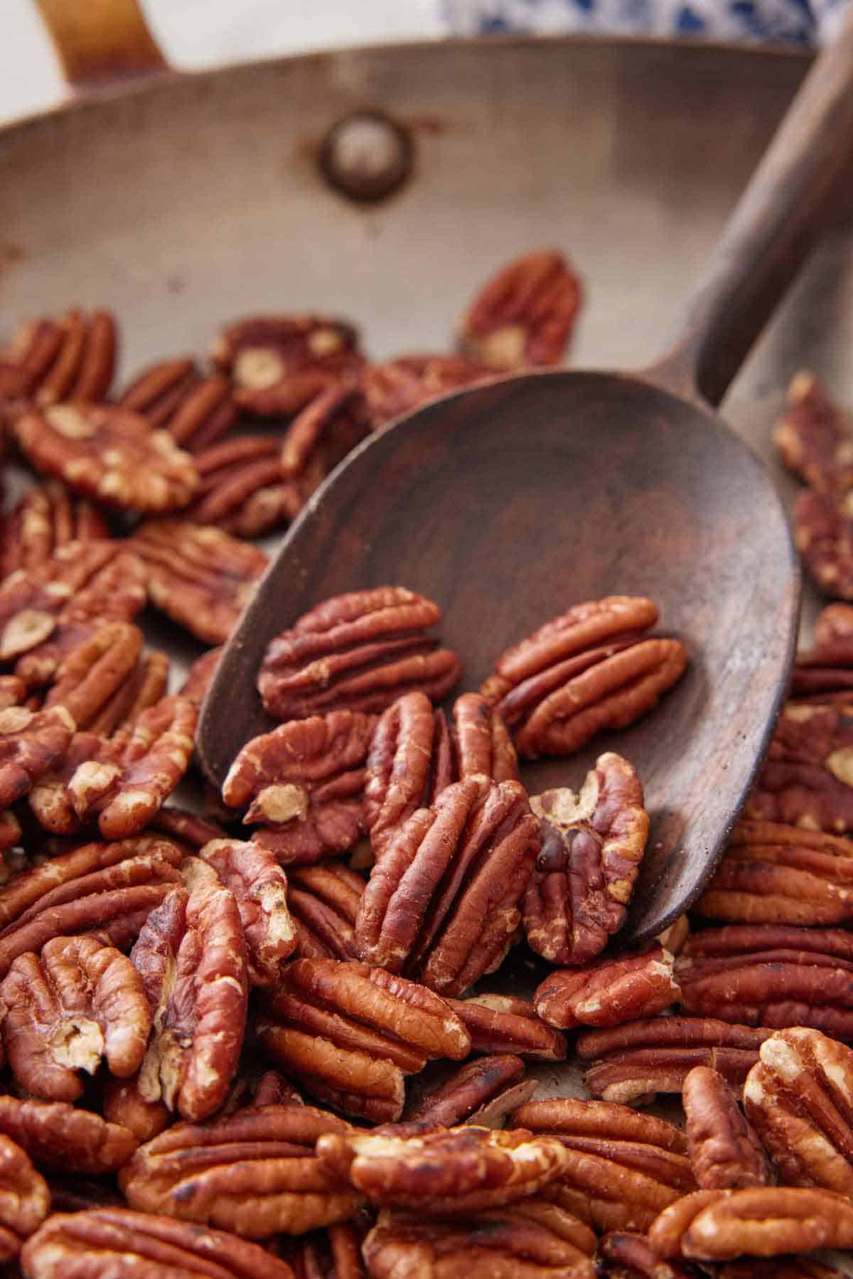 A wooden spoon scooping up some toasted pecans in a skillet.