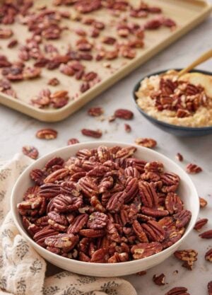 A bowl of toasted pecans with a sheet pan in the background and more pecans in a bowl over oatmeal.