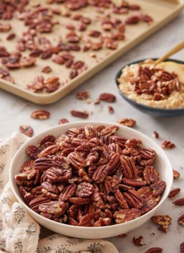 A bowl of toasted pecans with a sheet pan in the background and more pecans in a bowl over oatmeal.