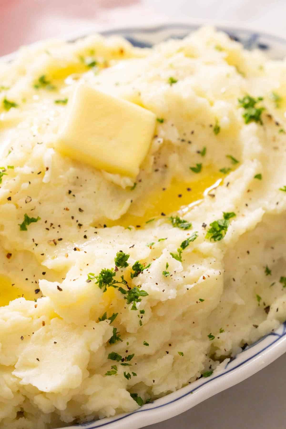 A close up view of mashed potatoes with a knob of softened butter on top along with chopped herbs.