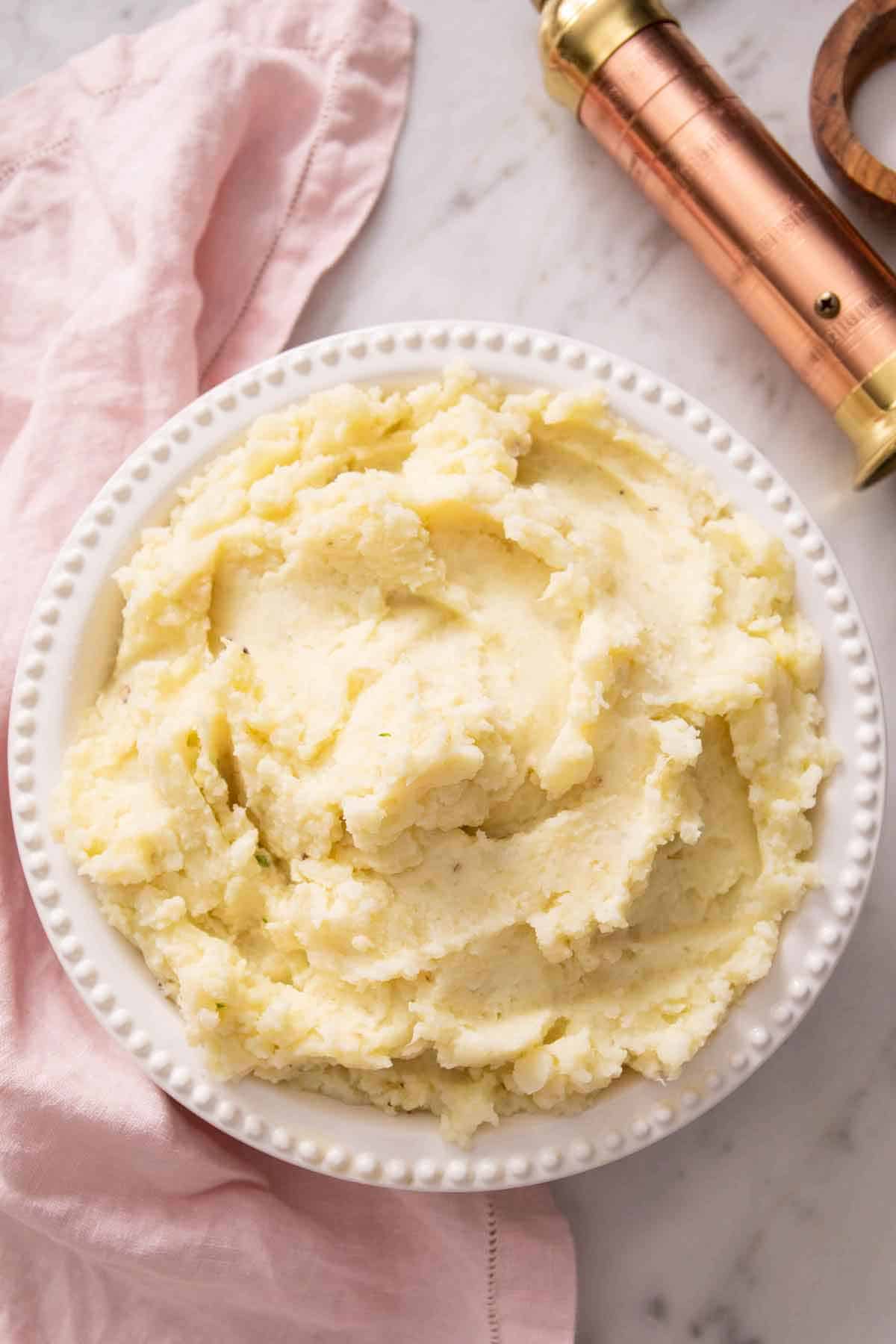 Overhead view of a bowl of mashed potatoes without garnishes and a pepper grinder off to the side.