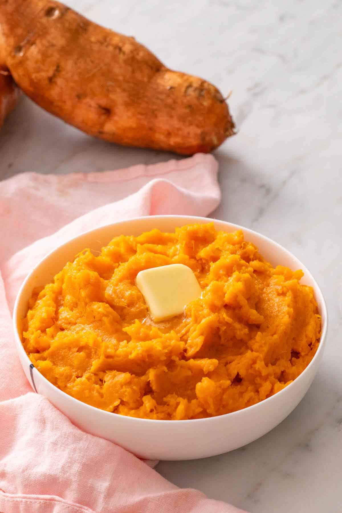 A bowl of mashed sweet potatoes with some slightly melted butter on top. A sweet potato in the background.