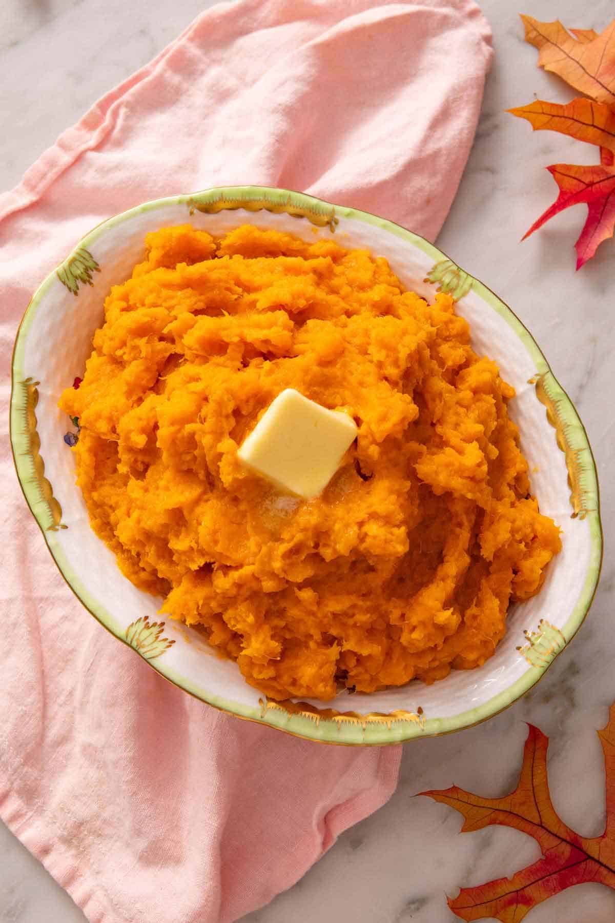 Overhead view of an oval platter of mashed sweet potatoes with butter on top. A pink linen underneath the platter and some leaves on the side.