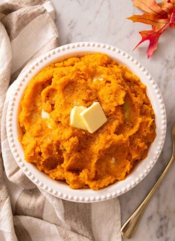 Overhead view of a bowl of mashed sweet potatoes with knobs of butter on top. Linen napkin and some leaves on the side.