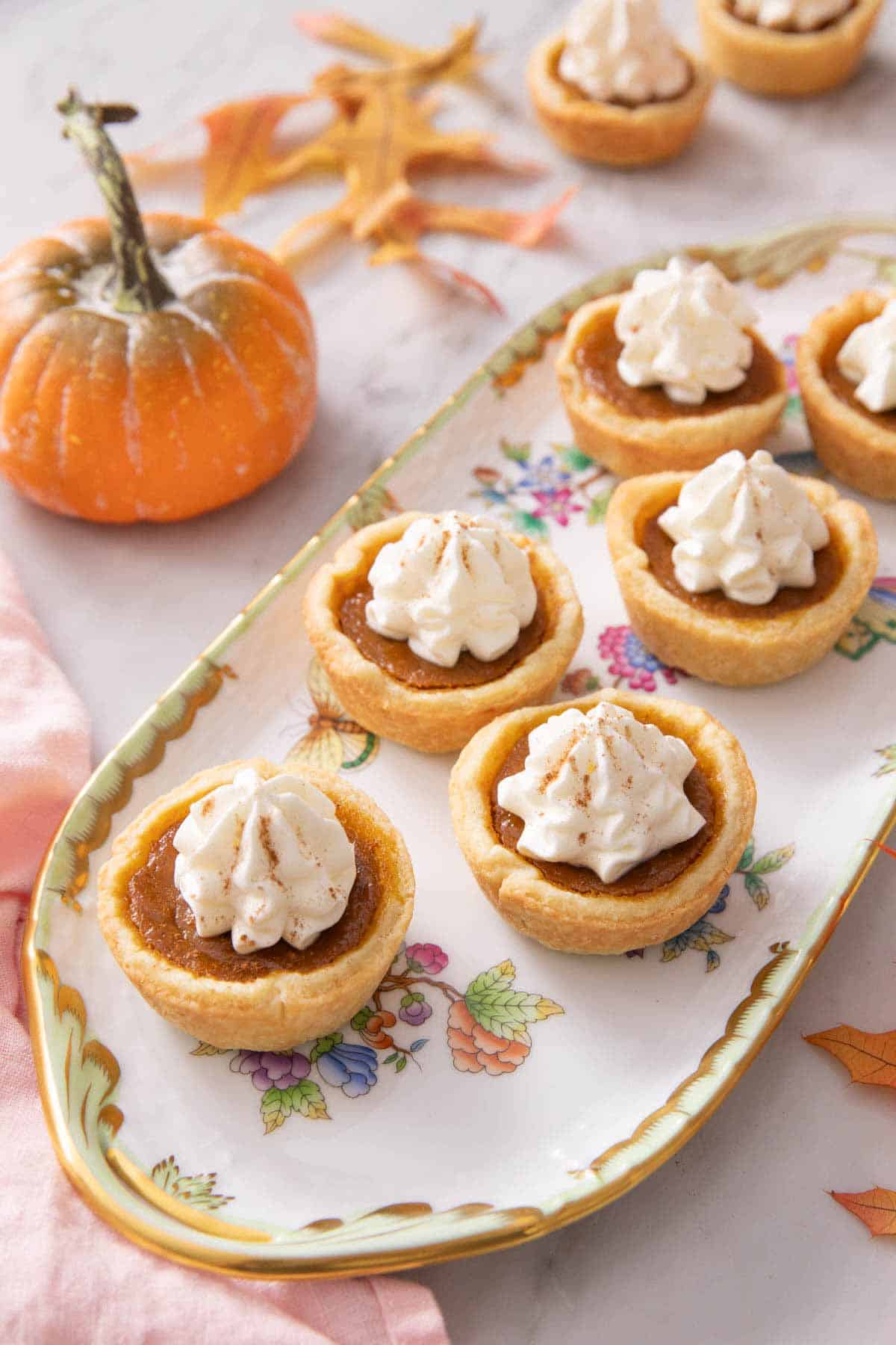A platter of mini pumpkin pies with whipped cream on top. A mini pumpkin and some leaves in the background.