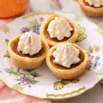 A plate with three mini pumpkin pies with more in the background. All topped with whipped cream.